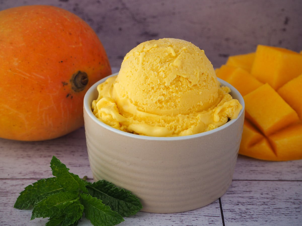 Mango ice cream in a small bowl, with fresh mint on the side and fresh mangoes in the background.