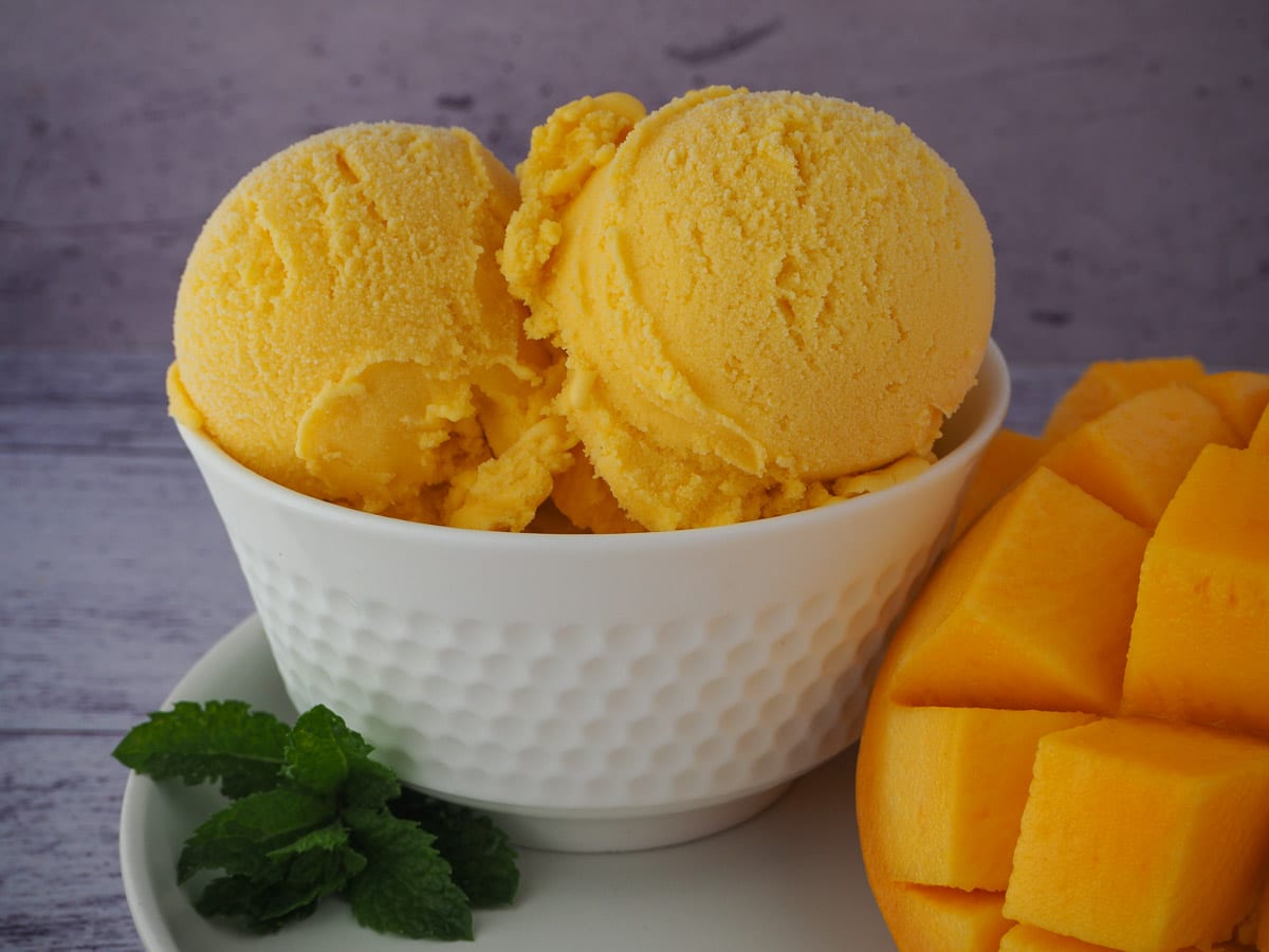 Scoops of mango ice cream in a white bowl, on a white plate with a spring of fresh mint and fresh mango.