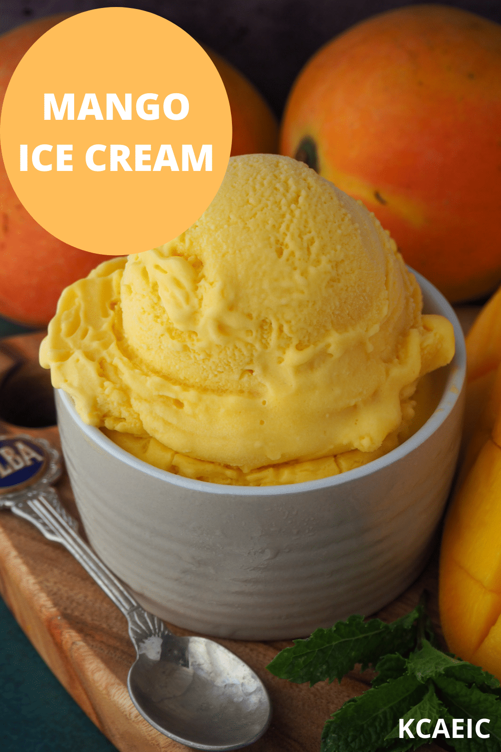 Mango ice cream in a small bowl, on a chopping board with a vintage spoon and mint in the foreground, and fresh mangos in the background and text overlay, mango ice cream, KCAEIC.