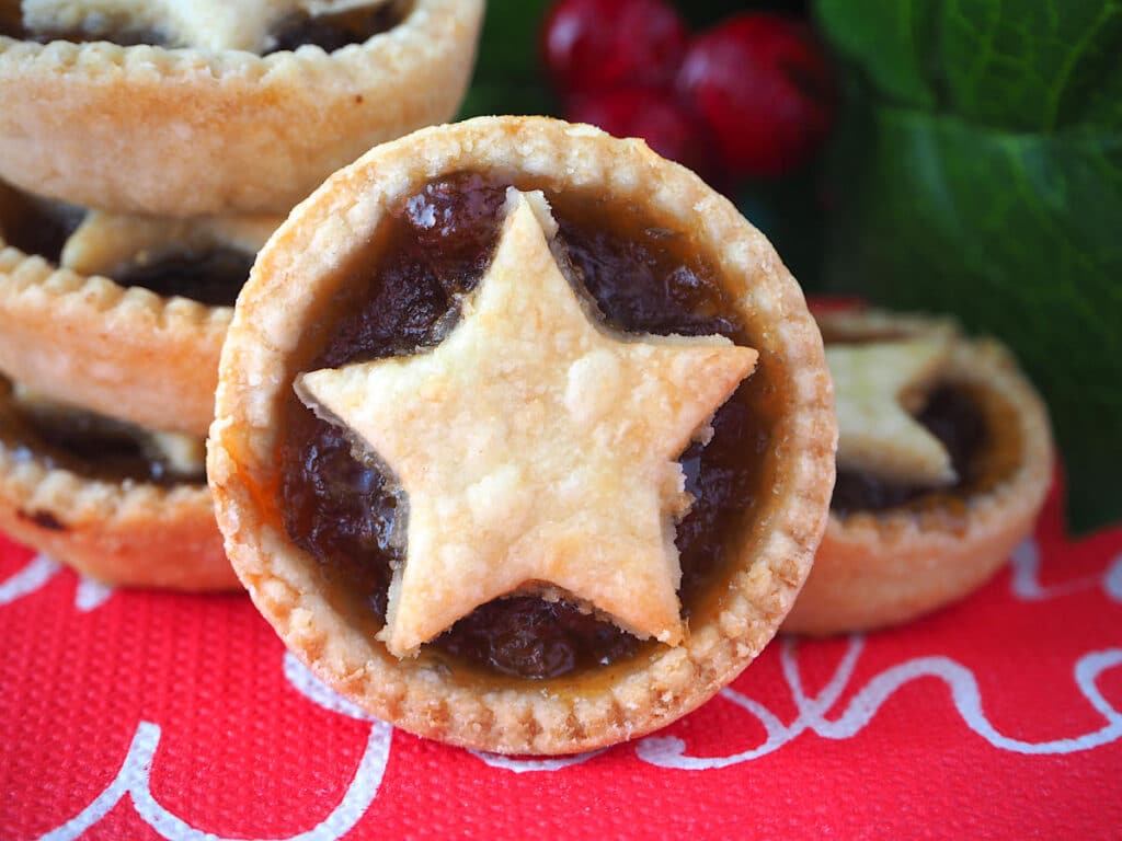 Mincemeat tart propped against a stack of mincemeat tarts, with holly in the background.