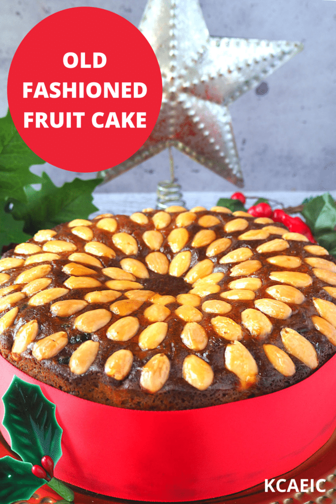 Old fashioned fruit cake decorated with almonds, with a red ribbon wrapped around it, on a gold plate, with holly decorations and a Christmas star in the background and text overlay, old fashioned fruit cake and KCAEIC.