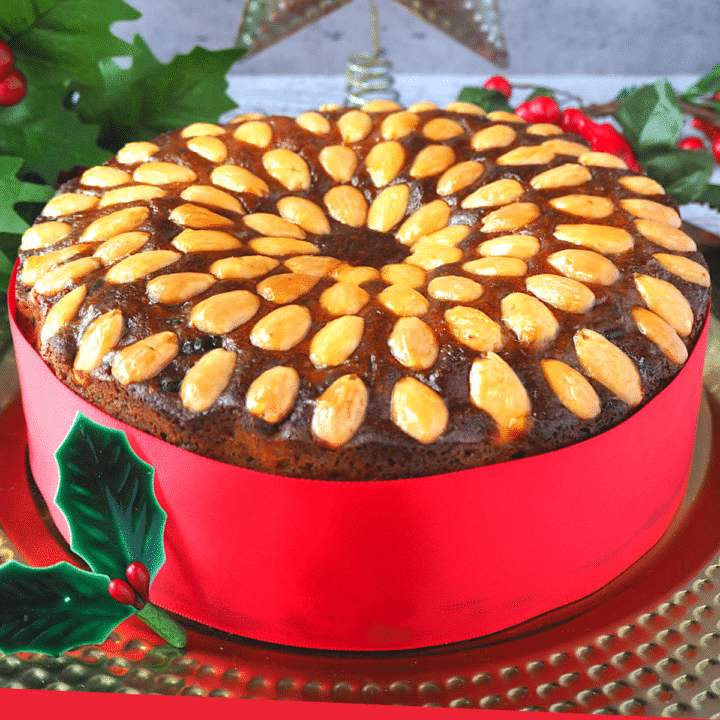 Old fashioned fruit cake decorated with almonds, with a red ribbon wrapped around it, on a gold plate, with holly decorations and a Christmas star in the background.