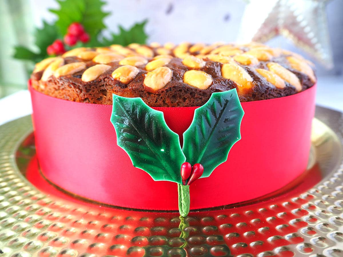 Old fashioned fruit cake decorated with almonds, with a red ribbon wrapped around it, on a gold plate, with holly decorations and a Christmas star in the background.