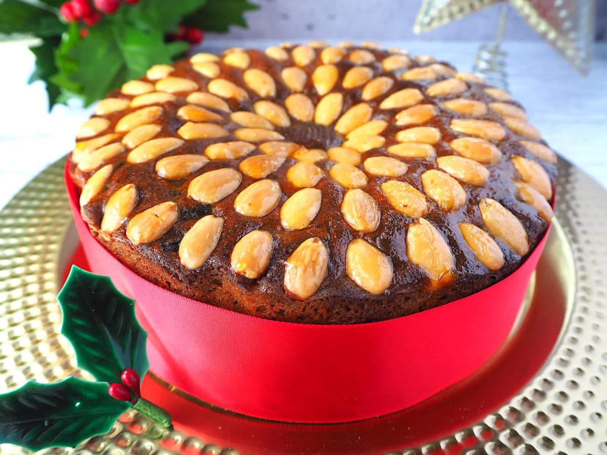 Old fashioned fruit cake decorated with almonds, with a red ribbon wrapped around it, on a gold plate, with holly decorations and a Christmas star in the background.