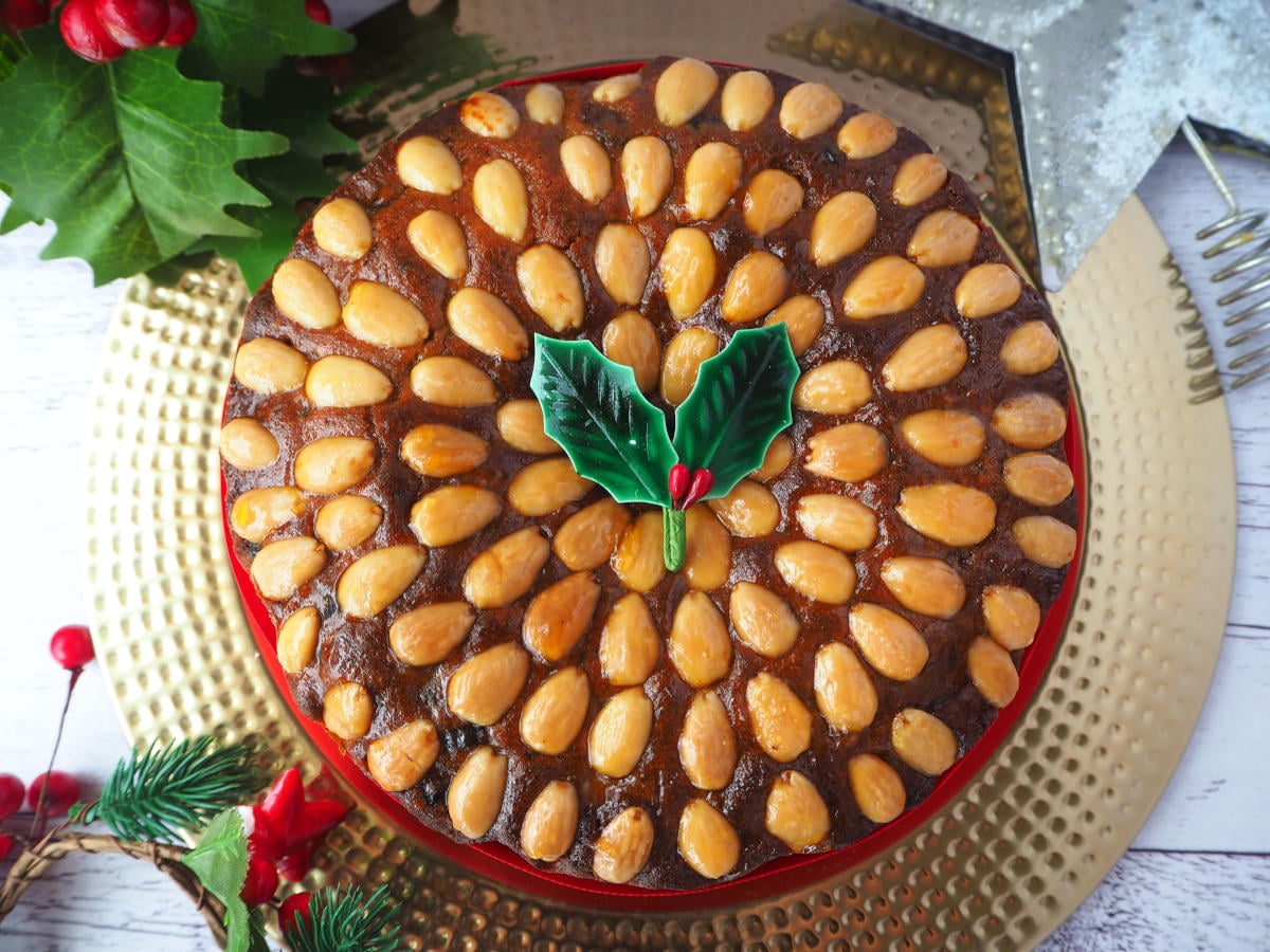 Top down view of old fashioned fruit cake decorated with almonds with a sprig of holly on top, with a red ribbon wrapped around it, on a gold plate, with holly decorations and a Christmas star on the sides.