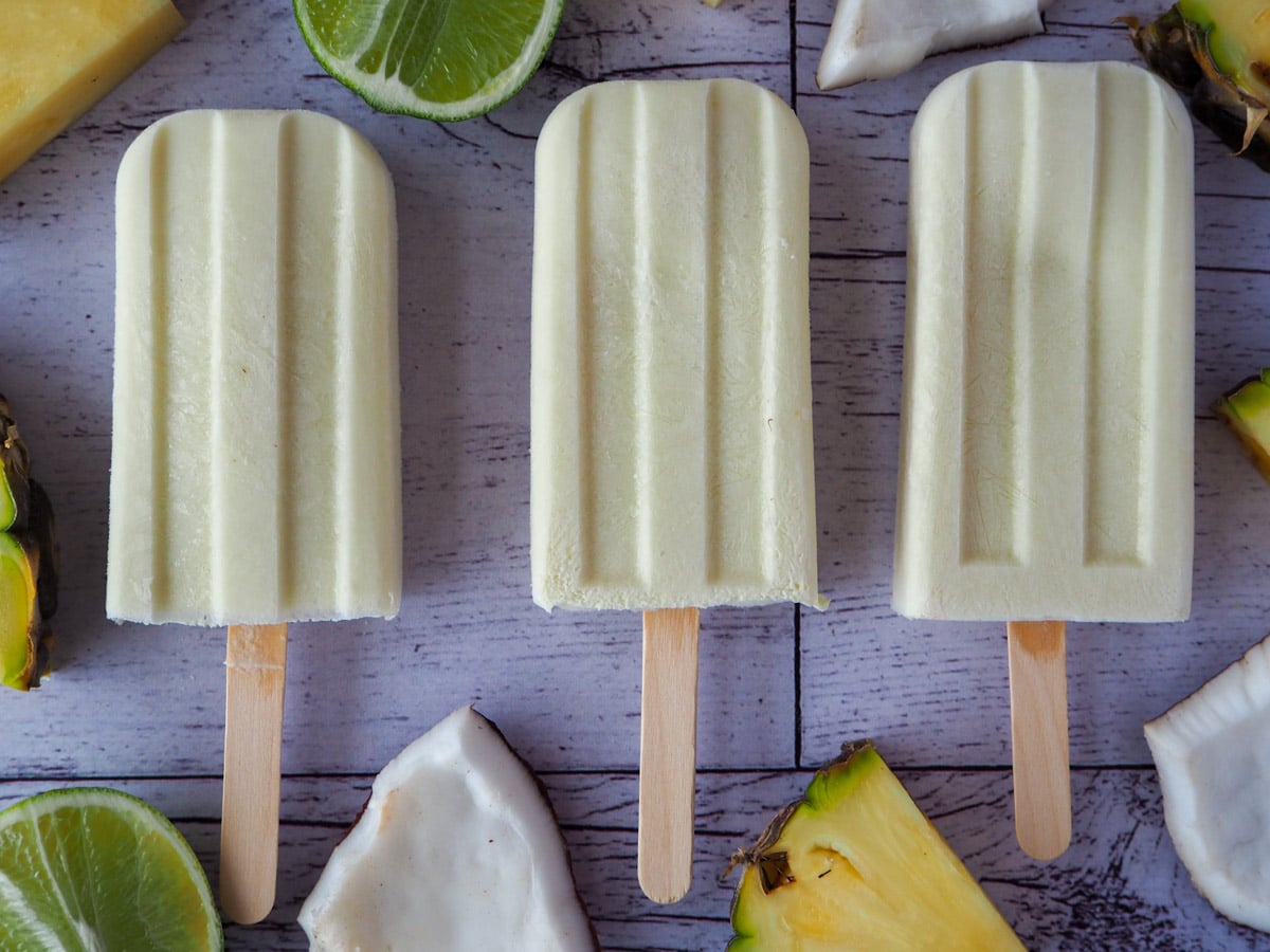 Row of three pina colada popsicles surrounded by fresh pineapple, coconut and limes.