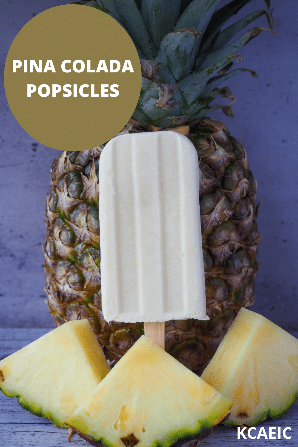 Single pina colada popsicle in front of a fresh pineapple, with fresh sliced pineapple in front and text overlay, pina colada popsicles and KCAEIC.