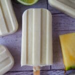 Close up of single pino colada popsicle, surrounded by other popsicles and fresh pineapple and lime.