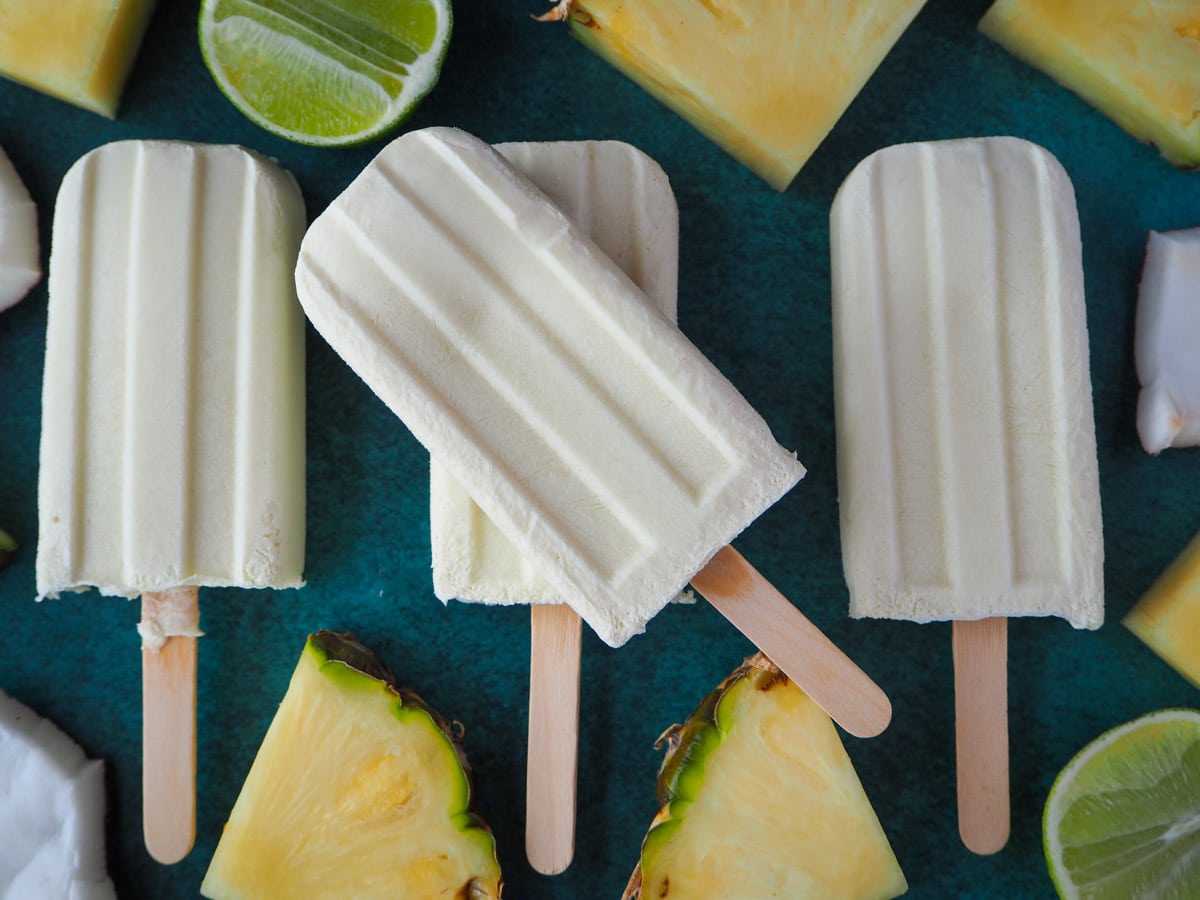 Row of three pina cola popsicles with another one stack on top in the middle, surrounded by fresh pineapple pieces, coconut and limes.