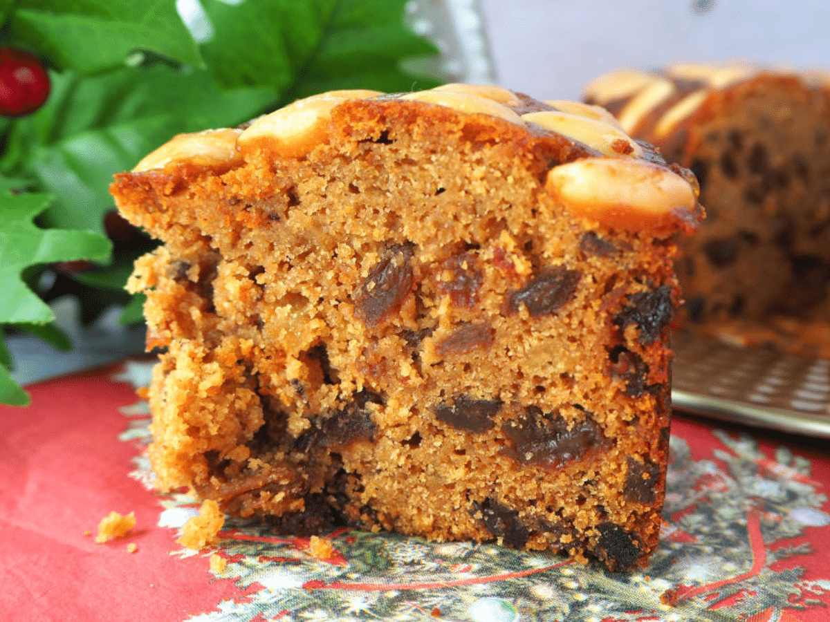 Side view of slice of old fashioned fruit cake, on a Christmas serviette, with holly and cut cake in the background.