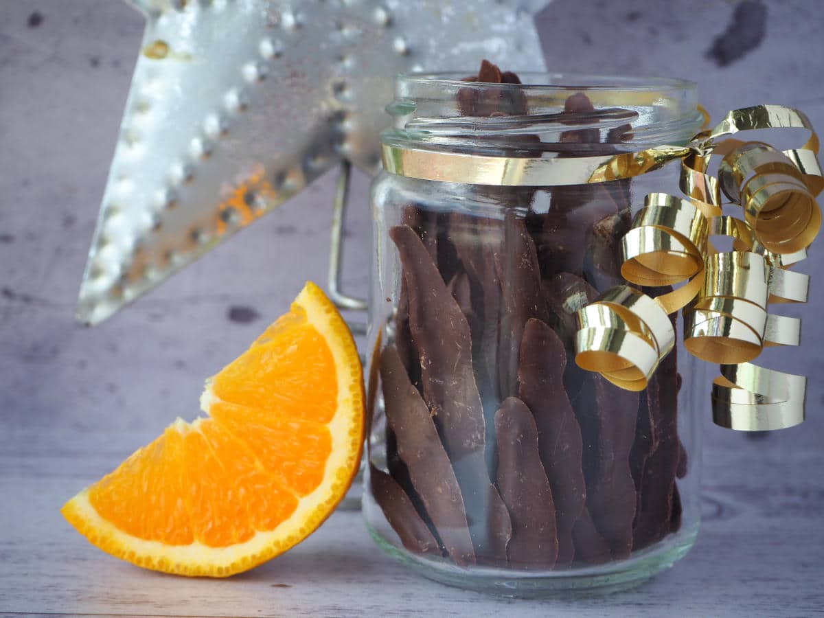 Glass jar of chocolate covered orange peel, with gold ribbon wrapped around it, slice of fresh orange leading against jar and Christmas star in background.