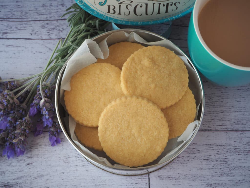 top down view gluten free shortbread in a biscuit tin, with a cup of tea and fresh lavender on the side.