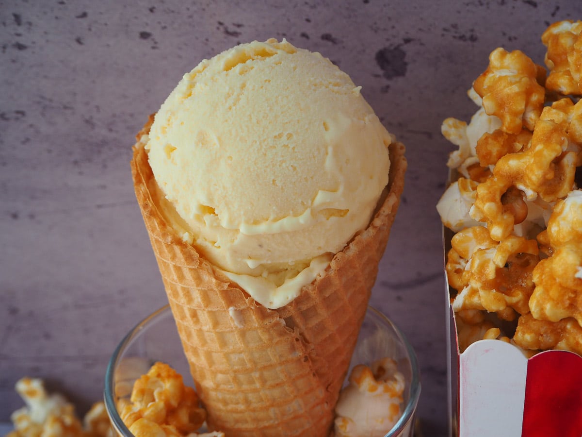 Popcorn ice cream in a waffle cone, in a glass filled with caramel popcorn, next to a box filled with caramel popcorn.