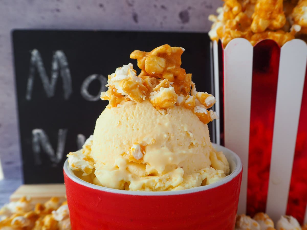 Popcorn ice cream in a red bowl, toped with caramel popcorn, next to a movie box filled with caramel popcorn, in front of a movie night blackboard.