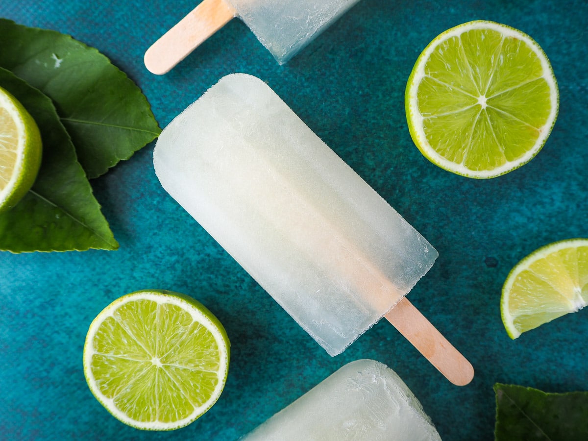 Lime popsicles surrounded by fresh cut limes, on a green background.
