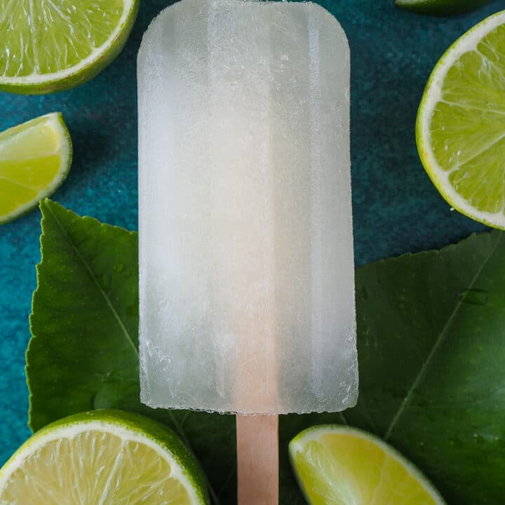 Single lime popsicle surrounded by fresh limes and leaves.