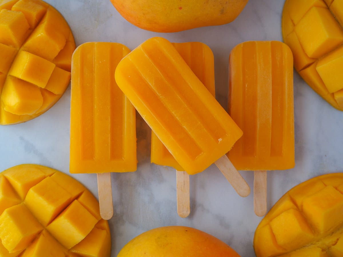 Row of three mango popsicles, with one stacked on top in the middle, surrounded by fresh mangos.