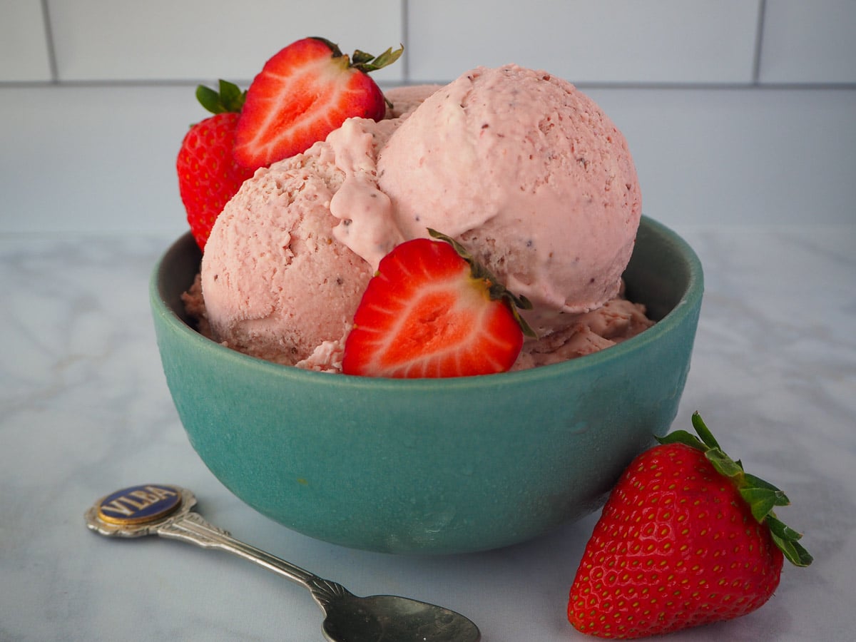 Scoop of no churn strawberry ice cream in a green bowl, with spoon on the side and fresh strawberries in the background.