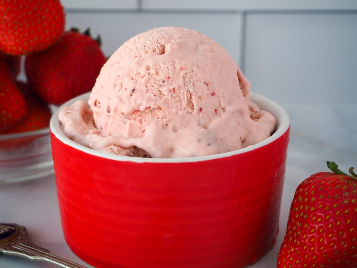 Scoop of no churn strawberry ice cream in a red bowl, with spoon on the side and fresh strawberries in the background.