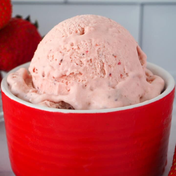 Close up of scoop of no churn strawberry ice cream in a red bowl with fresh strawberries in the background.