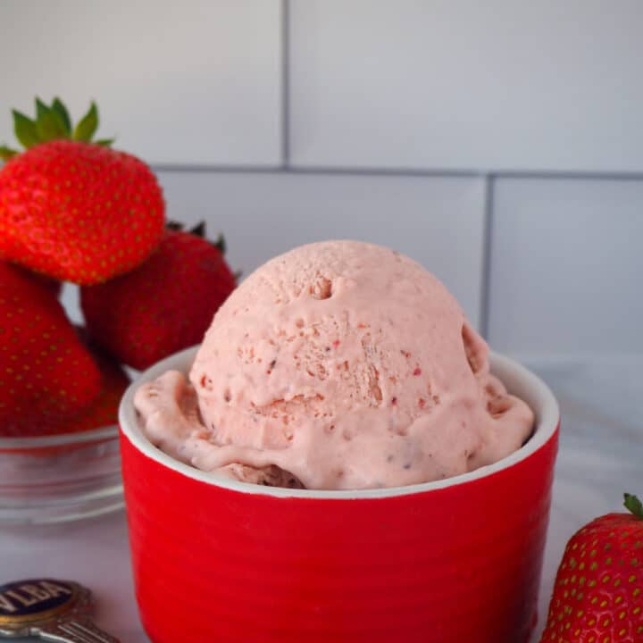 Scoop of no churn strawberry ice cream in a red bowl, with fresh strawberries and vintage spoon on the side.