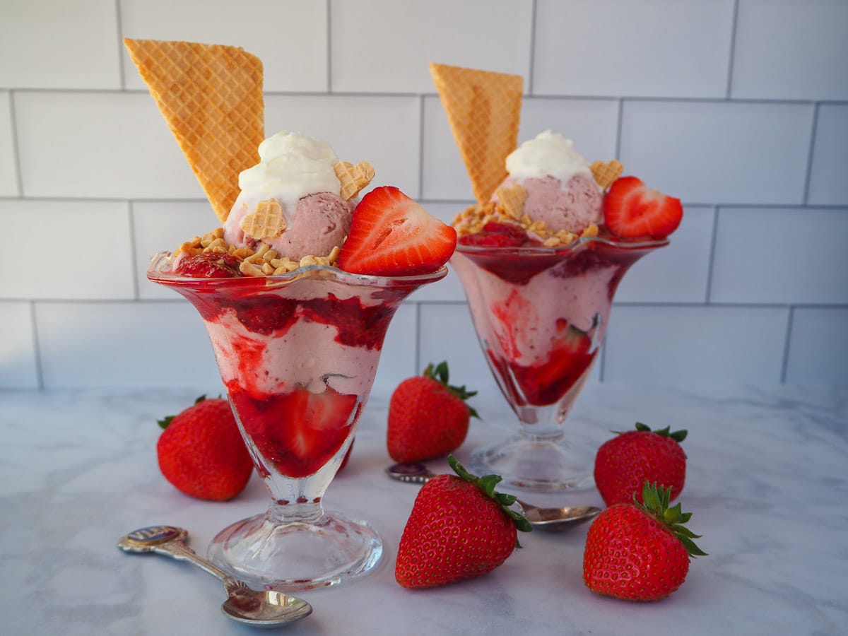 Strawberry sundae with another sundae in the background, surrounded by fresh strawberries and two vintage spoons.