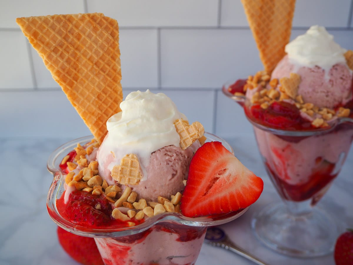 Strawberry sundae with another sundae in the background, surrounded by fresh strawberries and two vintage spoons.