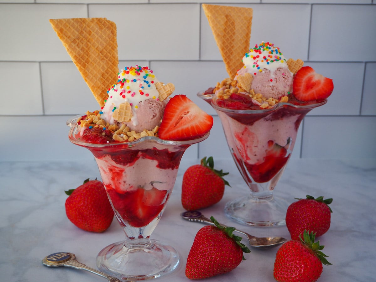 Two strawberry sundaes with strawberry ice cream, strawberry sauce, fresh strawberry, whipped cream, nuts, waffles and rainbow sprinkles, surrounded by fresh strawberries and two vintage spoons.
