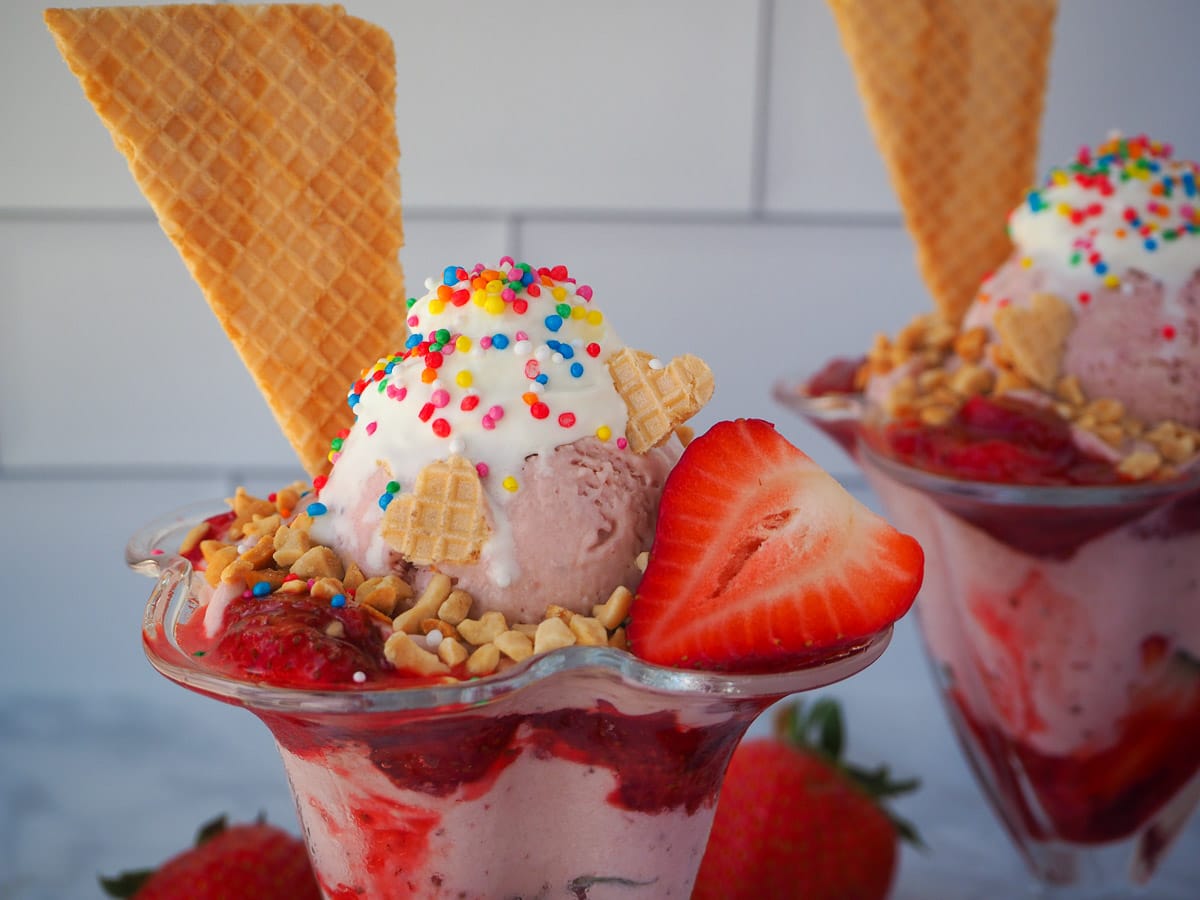Close up of strawberry sundae with strawberry ice cream, strawberry sauce, fresh strawberry, whipped cream, nuts, waffles and rainbow sprinkles, with a second sundae in the background, surrounded by fresh strawberries.