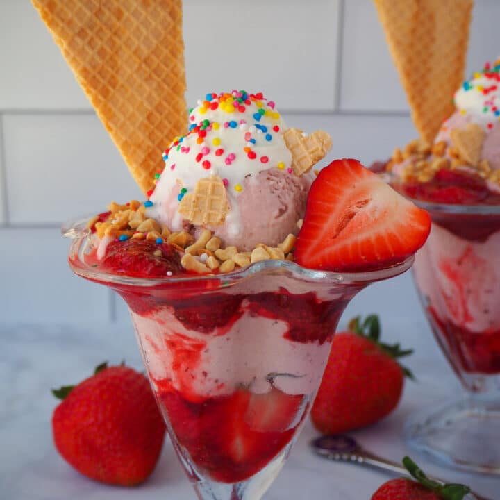 Close up of strawberry sundae with strawberry ice cream, strawberry sauce, fresh strawberry, whipped cream, nuts, waffles and rainbow sprinkles, with a second sundae in the background, fresh strawberries around them and vintage spoons.