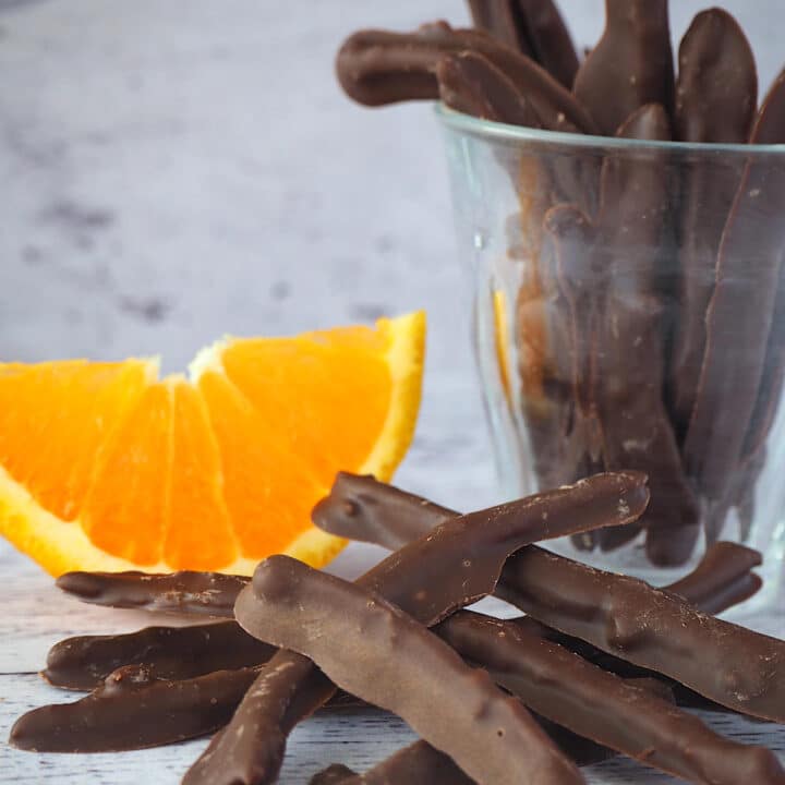 Chocolate covered orange peel with with fresh orange in the back ground and glass of chocolate covered orange peel.
