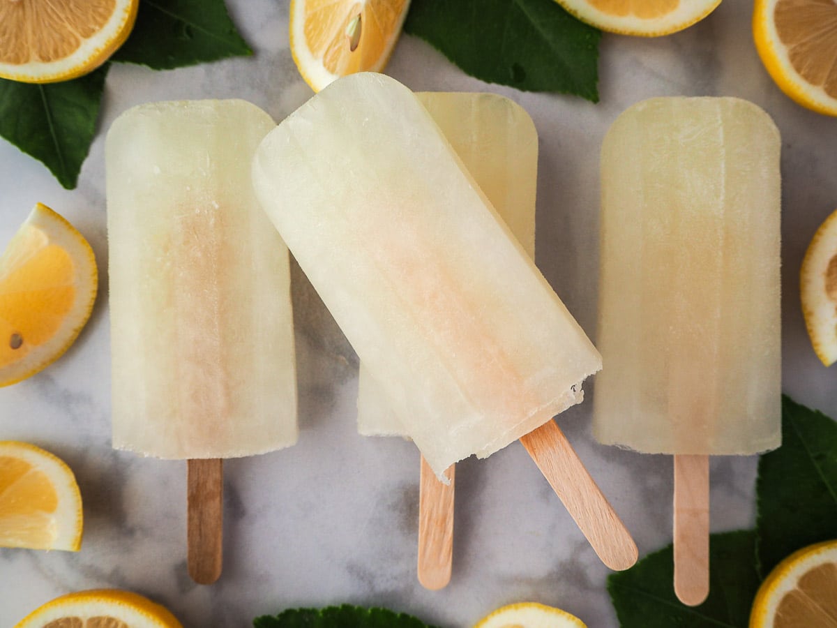 Row of three lemonade popsicles with a forth stacked on top, surrounded by fresh lemon slices and lemon leaves.