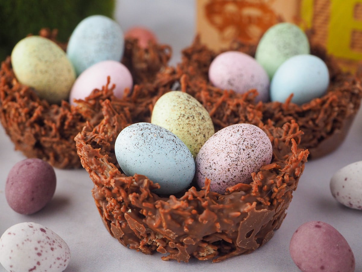 Group of three shredded wheat nests with extra speckled Easter eggs, Easter themed blocks and green bunny figurine in the background.