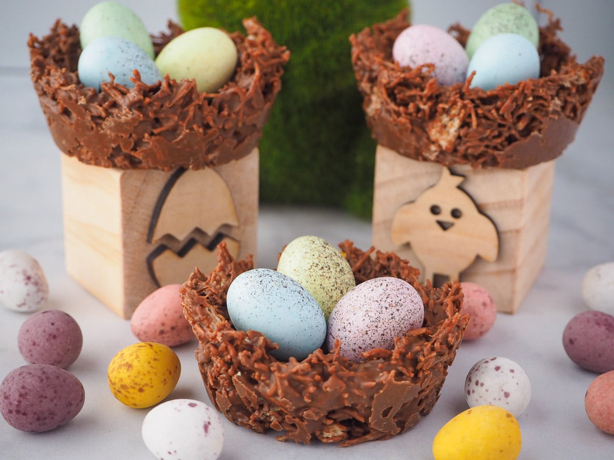 Group of three shredded wheat nests, surrounded by extra speckled Easter eggs, with two nests on Easter themed blocks and a green bunny figurine in the middle.