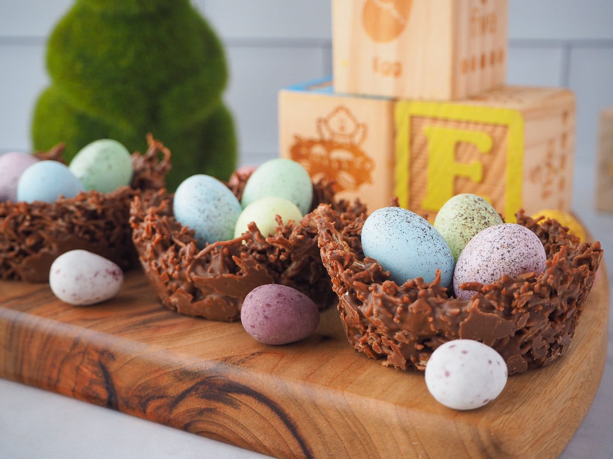 Row of three shredded wheat nests on a board, with extra speckled Easter eggs and Easter themed blocks and green bunny figurine in the background.