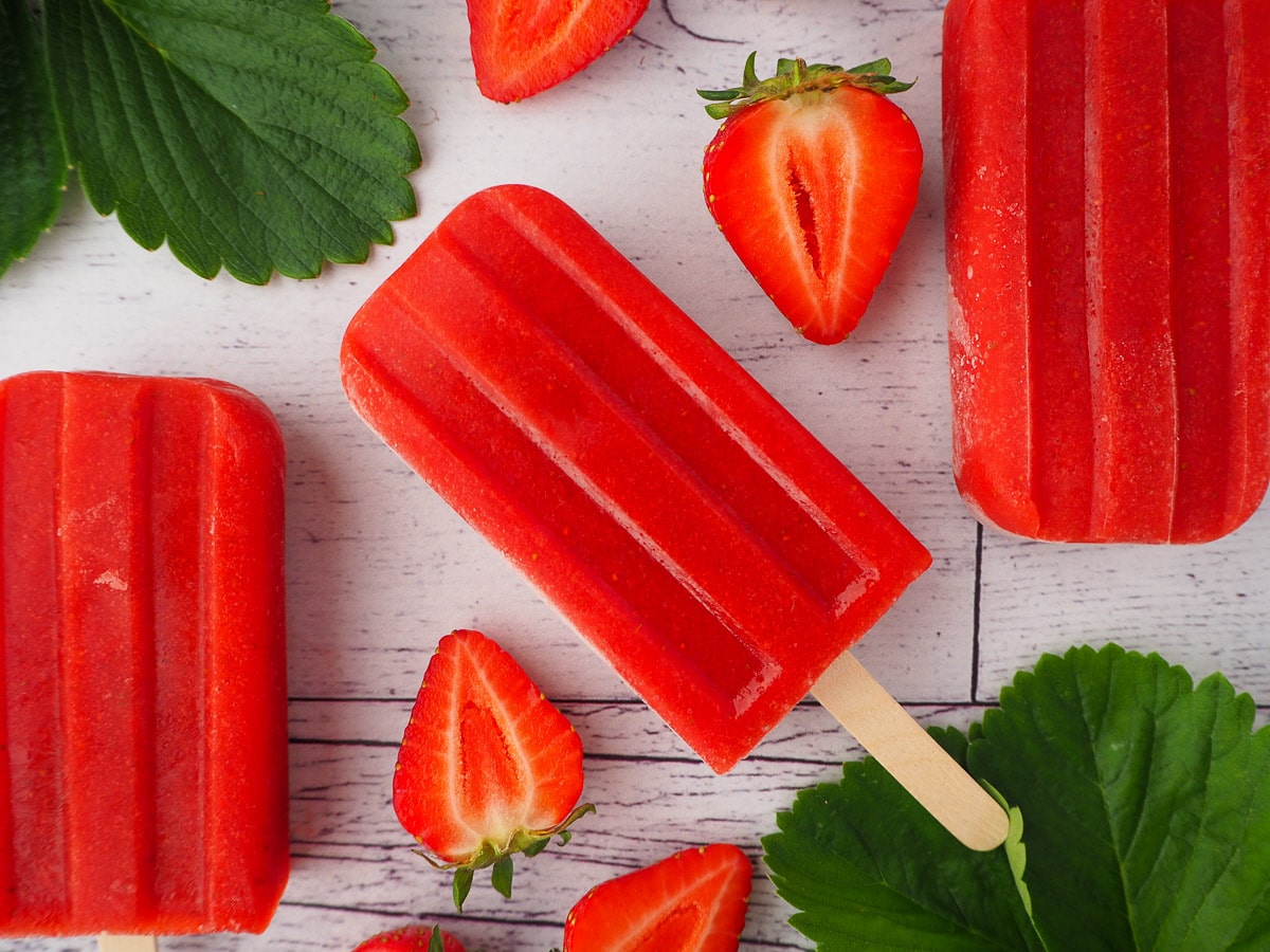 Row of three strawberry popsicles, surrounded by fresh strawberries and strawberry leaves.