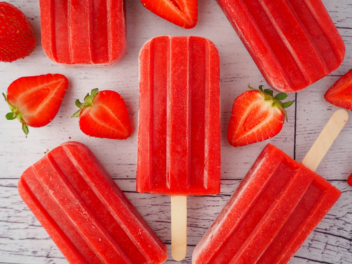 Close up of a strawberry popsicle, surrounded by fresh strawberries and other strawberry popsicles.