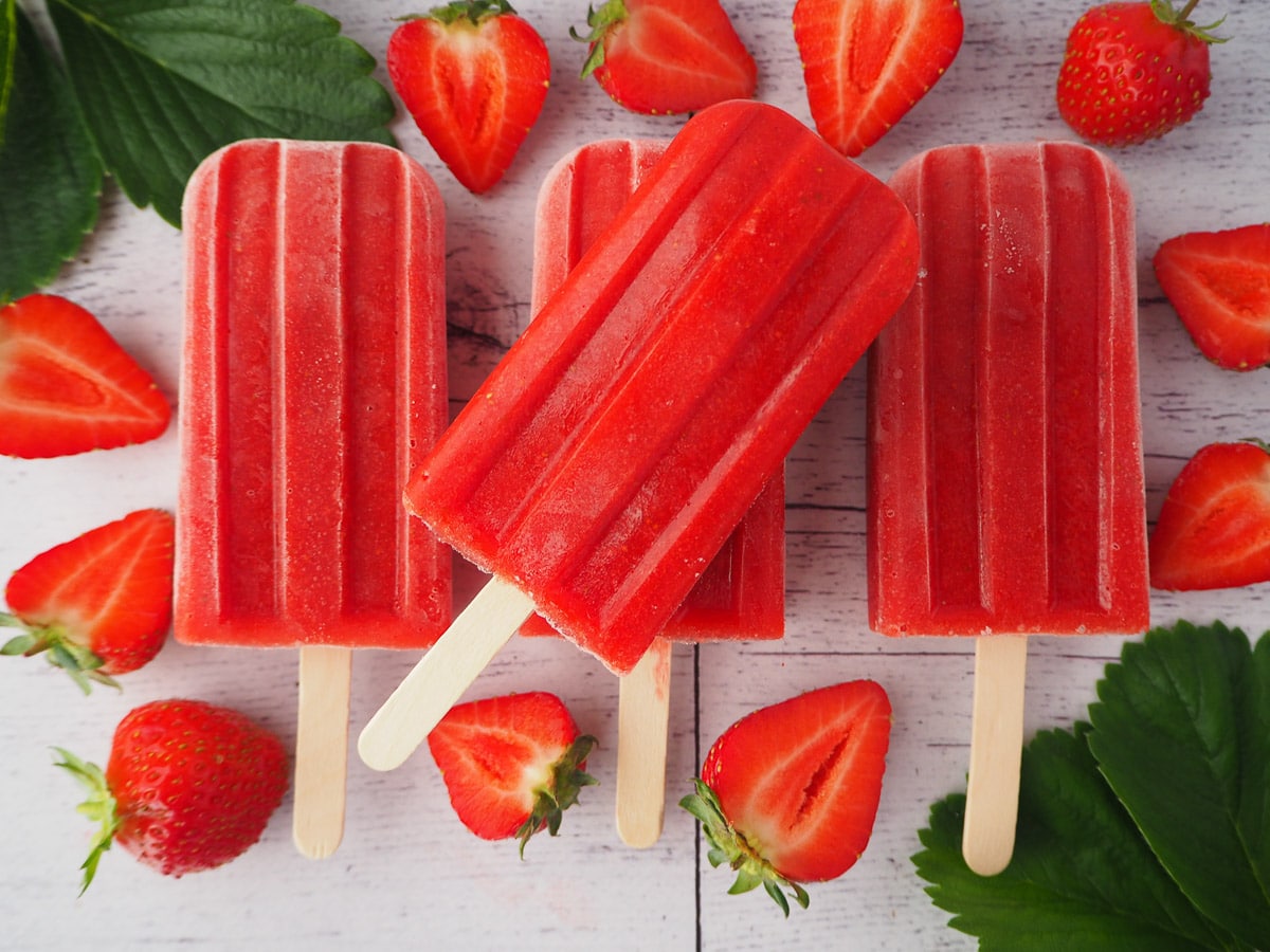 Row of three strawberry popsicles with a 4th stacked on top, surrounded by fresh strawberries and strawberry leaves.