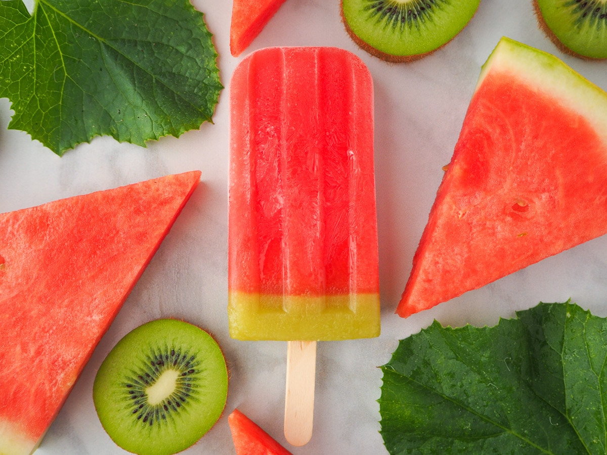 Single watermelon ice lolly surrounded by fresh watermelon, watermelon leaves and kiwi fruit.