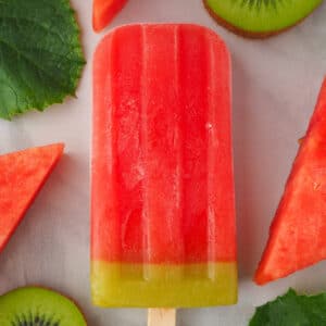 Single watermelon ice lolly surrounded by fresh watermelon, watermelon leaves and kiwi fruit.