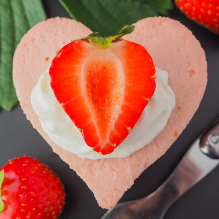 Close up heart shaped mini strawberry cheesecake with whipped cream, fresh strawberries, fresh strawberry leaves and a vintage fork.