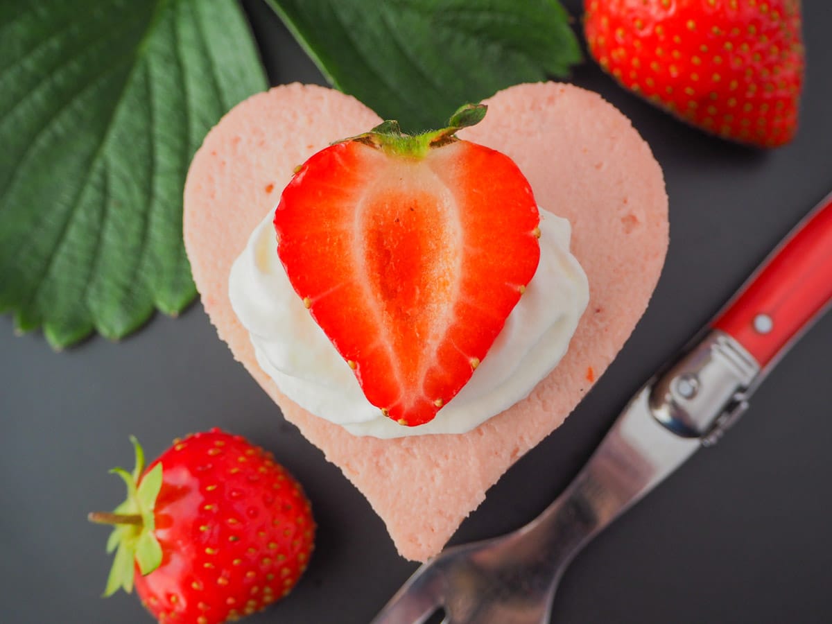 Heart shaped mini strawberry cheesecake with whipped cream, fresh strawberries, fresh strawberry leaves and a vintage fork.