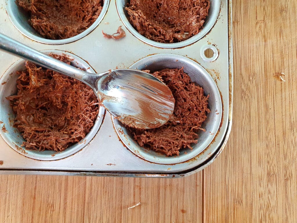 Shaping shredded wheat and chocolate mix into nest shapes in muffin tins with a spoon.