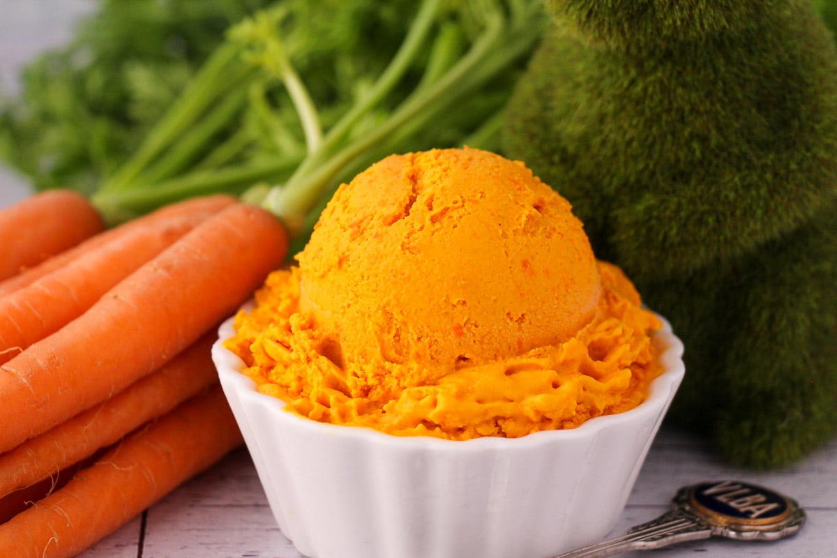 Scoop of carrot ice cream in a white bowl, with a vintage spoon and fresh Dutch carrots on the side, a green grass rabbit figure.