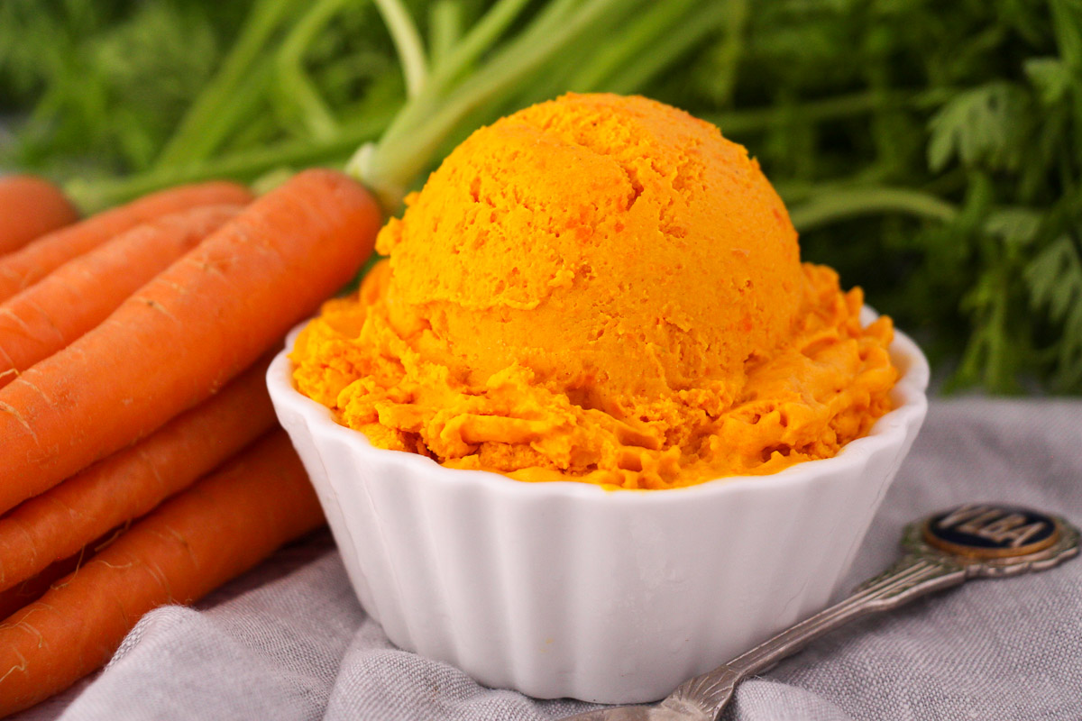 Scoop of carrot ice cream in a white bowl, with a vintage spoon and fresh Dutch carrots on the side.