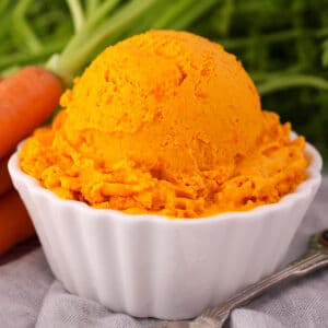 Close ups scoop of carrot ice cream in a white bowl, with a vintage spoon and fresh Dutch carrots on the side.