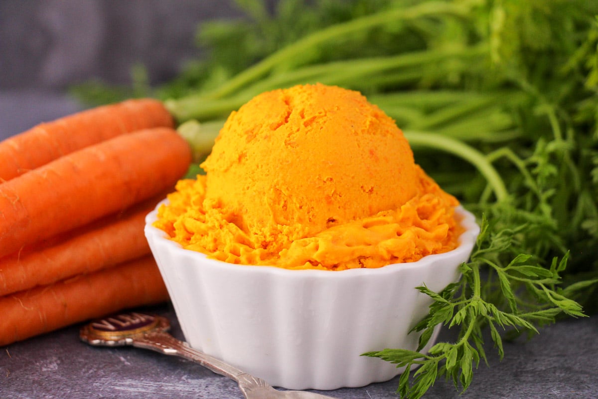 Scoop of carrot ice cream in a white bowl, with a vintage spoon and fresh Dutch carrots on the side.