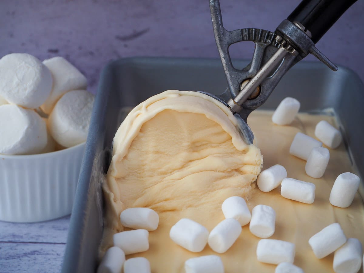 Scooping marshmallow ice cream, with mini marshmallows on top and bowl of marshmallows in the background.