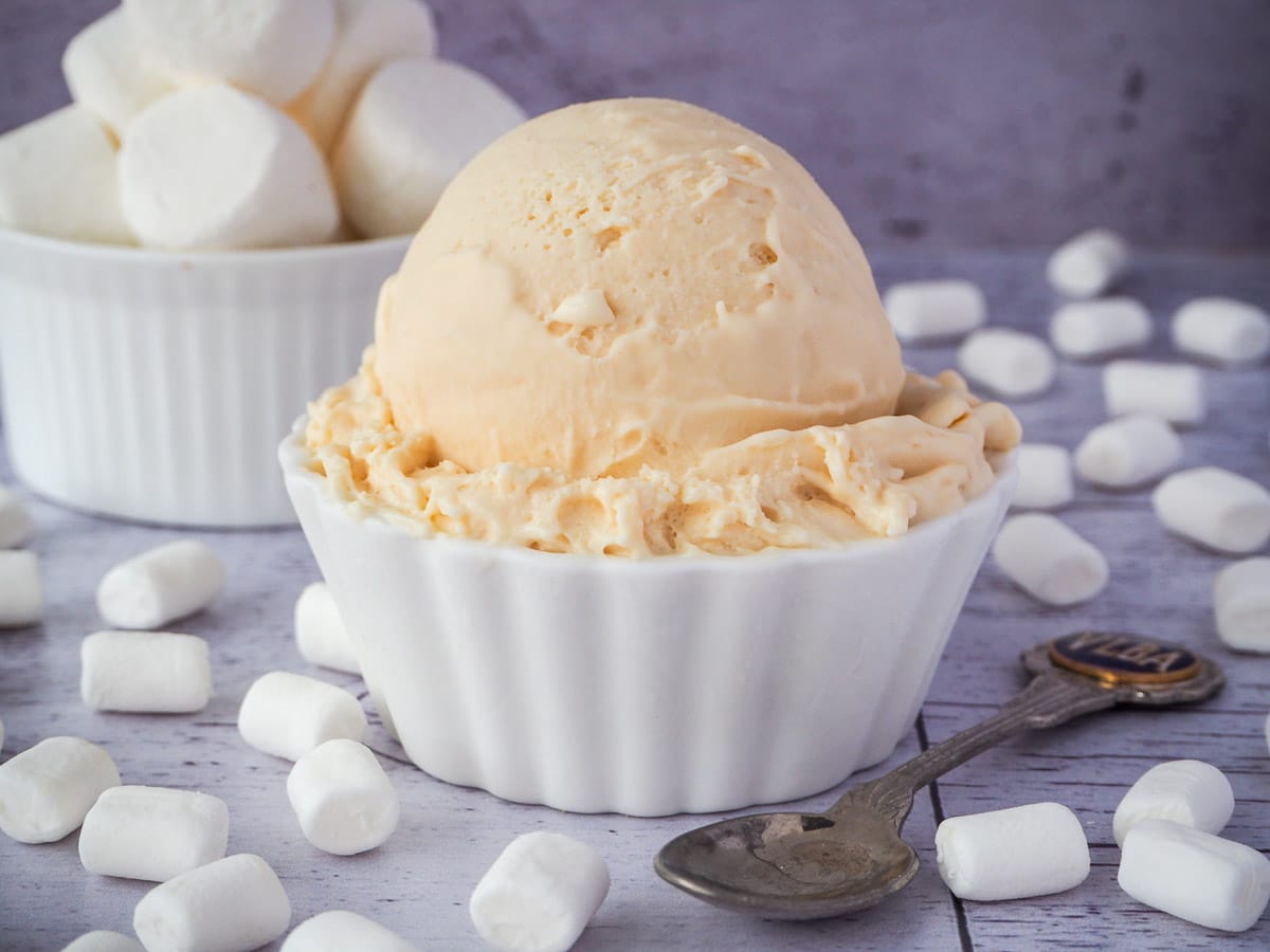 Scoop of marshmallow ice cream in a decorative white bowl, with vintage spoon on the side, with a bowl of marshmallows in the background, surrounded by mini marshmallows.