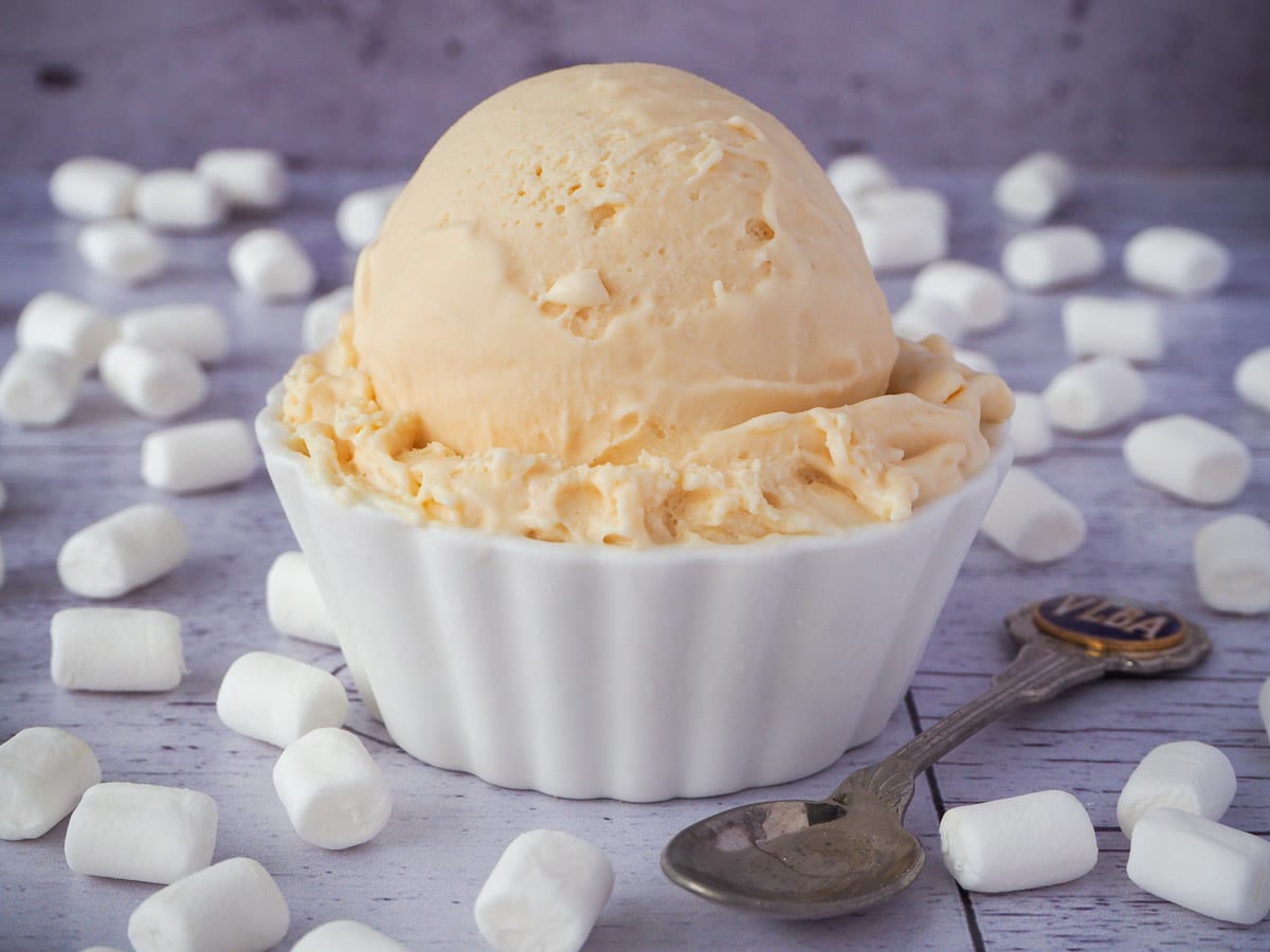 Scoop of marshmallow ice cream in a decorative white bowl, with vintage spoon on the side, surrounded by mini marshmallows.